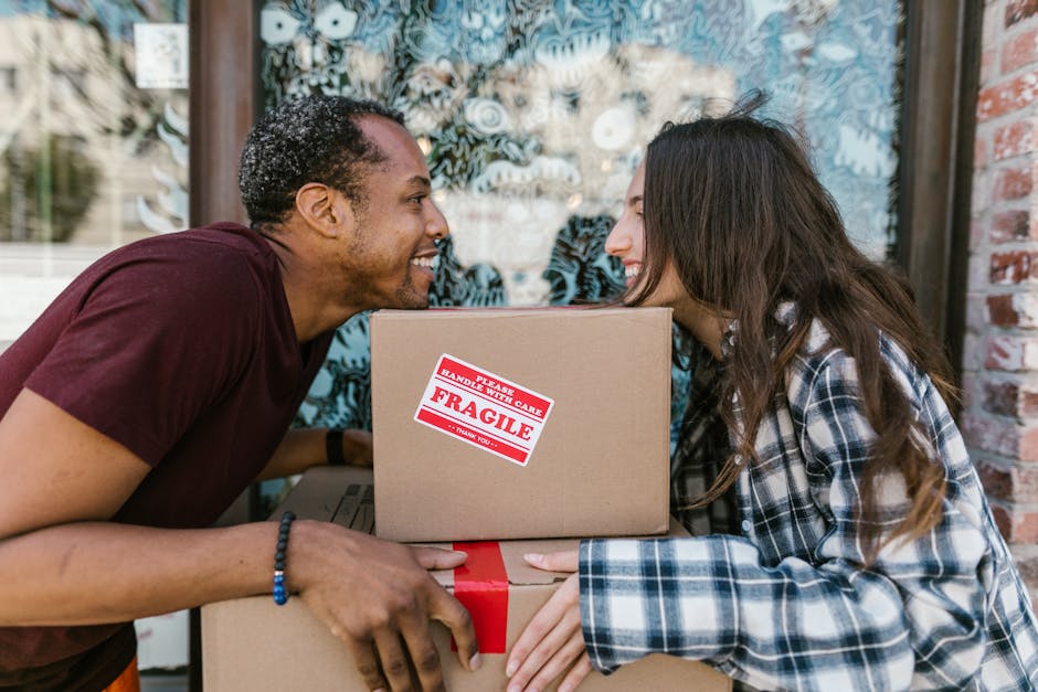 A man and woman with light skin are engaged in a home relocation process, smiling and facing each other as they handle cardboard boxes. The man, wearing a maroon t-shirt and a beaded bracelet, is perched outside a brick property and is holding a medium-sized cardboard box with a red and white 'FRAGILE' sign, indicating careful handling during packing and moving. The woman, dressed in a plaid shirt, is also holding and supporting the same box, and their foreheads are touching in a friendly, cheerful manner, suggesting teamwork during furniture transport. Behind them, a large window with a decorative window film and some greenery are visible, with natural daylight illuminating the scene. The scene exemplifies the loading process during a home relocation, with materials such as cardboard boxes, protective packaging, and possible use of hand-held lifting techniques supported by the presence of multiple boxes. This image reflects professional removals services provided by Ealing Man and Van, emphasizing the careful handling and packing involved in house removals and household moves.