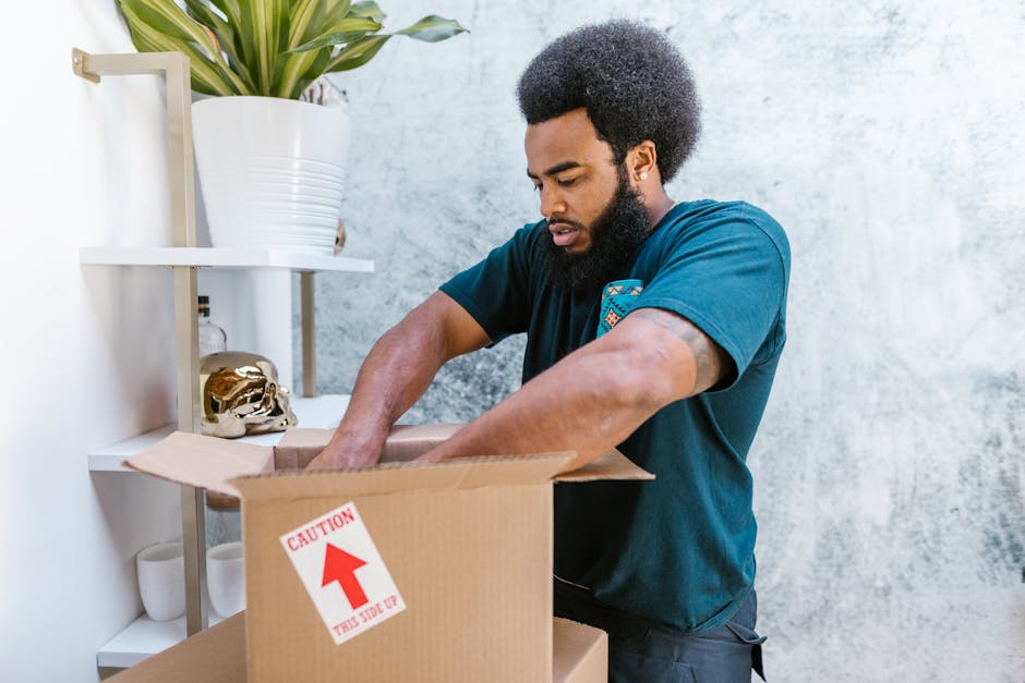 A man with a dark beard and curly hair, wearing a blue T-shirt, is shown packing items into a cardboard box during a home relocation process. He is standing indoors next to a white shelving unit that holds a large potted plant, a golden decorative skull, and various small objects. The man’s hands are inside the open box, which is on a table or countertop, and his focused expression indicates careful packing. The cardboard box has a red and white CAUTION label with an arrow pointing upward, emphasizing safe handling and proper packing techniques. The environment features a plain light-textured wall, and natural lighting illuminates the scene, reflecting an organized and methodical approach to furniture transport and packing in a house move. Such packing procedures are typically part of services offered by companies like Ealing Man and Van for residential removals and moving logistics, supporting efficient furniture transport and home relocation activities.