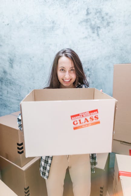 A man and woman with light skin are engaged in a home relocation process, smiling and facing each other as they handle cardboard boxes. The man, wearing a maroon t-shirt and a beaded bracelet, is perched outside a brick property and is holding a medium-sized cardboard box with a red and white 'FRAGILE' sign, indicating careful handling during packing and moving. The woman, dressed in a plaid shirt, is also holding and supporting the same box, and their foreheads are touching in a friendly, cheerful manner, suggesting teamwork during furniture transport. Behind them, a large window with a decorative window film and some greenery are visible, with natural daylight illuminating the scene. The scene exemplifies the loading process during a home relocation, with materials such as cardboard boxes, protective packaging, and possible use of hand-held lifting techniques supported by the presence of multiple boxes. This image reflects professional removals services provided by Ealing Man and Van, emphasizing the careful handling and packing involved in house removals and household moves.