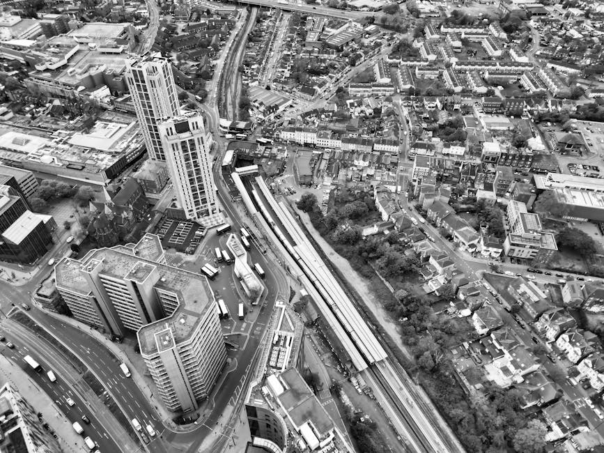 A black-and-white aerial photograph showing a dense urban area with high-rise buildings, residential houses, and a network of roads. Visible in the image are multiple multi-storey office and apartment buildings with flat roofs, some with rooftop amenities or equipment, situated near a major road junction. The roads are busy with various vehicles, including cars and buses, some of which are navigating curved and straight sections of the street. An elevated train track runs through the scene, with a train approaching or paused at a station platform adjacent to a row of lower residential buildings and houses. The surrounding streets are lined with trees and smaller structures, indicating a mixed-use neighbourhood. This urban landscape captures a typical environment where house removals and furniture transport services, such as those provided by Ealing Man and Van, might operate within a busy city setting near Ealing Broadway station, W5. The image emphasizes the complexity of city logistics involved in home relocation and moving logistics in such environments.