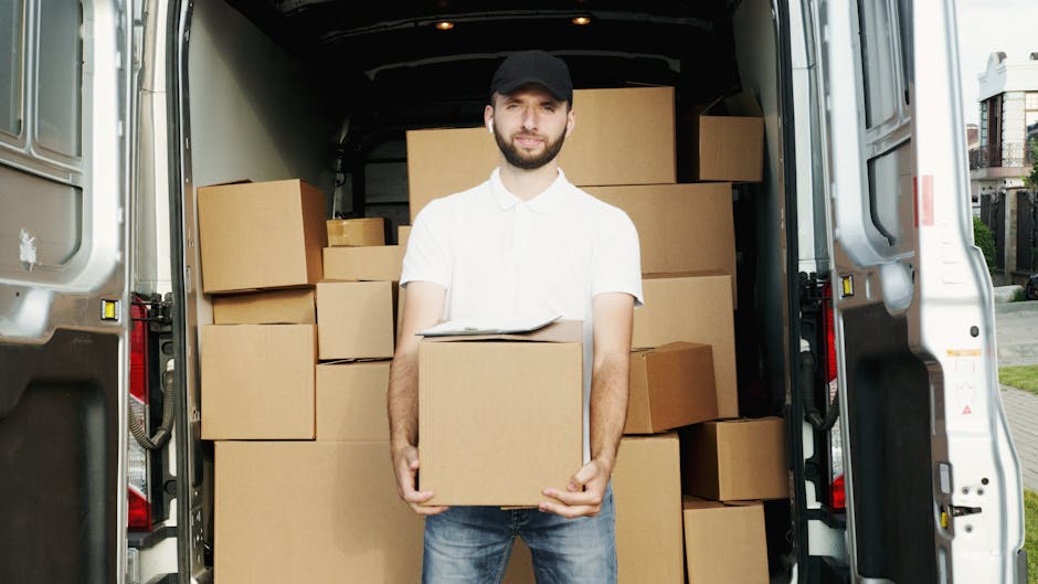 A man wearing a white shirt and black cap stands inside an open delivery van, holding a cardboard box and facing the camera. The van is filled with multiple cardboard boxes of various sizes, some wrapped in plastic or packing paper, stacked neatly inside the cargo area. The van is parked outdoors on a paved street with residential buildings visible in the background. The scene depicts a home relocation or furniture transport process, with a focus on the loading or unloading stage of a house removal. [COMPANY_NAME], specializing in removals, is involved in the logistics of this packing and moving operation, demonstrating the careful handling of items during a house move. The lighting is natural, highlighting the interior of the van and the boxes, emphasizing the logistics of tight access moves often encountered in property clearances or apartment relocations, consistent with the tips for tight access moves in Ealing across the [PAGE_TITLE].