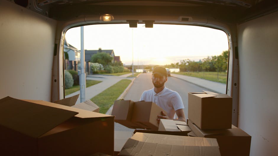 A man wearing a white shirt and black cap stands inside an open delivery van, holding a cardboard box and facing the camera. The van is filled with multiple cardboard boxes of various sizes, some wrapped in plastic or packing paper, stacked neatly inside the cargo area. The van is parked outdoors on a paved street with residential buildings visible in the background. The scene depicts a home relocation or furniture transport process, with a focus on the loading or unloading stage of a house removal. [COMPANY_NAME], specializing in removals, is involved in the logistics of this packing and moving operation, demonstrating the careful handling of items during a house move. The lighting is natural, highlighting the interior of the van and the boxes, emphasizing the logistics of tight access moves often encountered in property clearances or apartment relocations, consistent with the tips for tight access moves in Ealing across the [PAGE_TITLE].