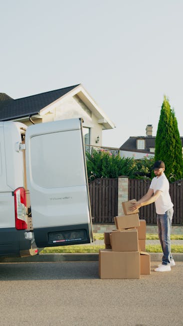 A male worker with a beard, wearing a white polo shirt and a black cap, standing outside near a white van during a home relocation process. He is holding a cardboard box, and several other boxes are s